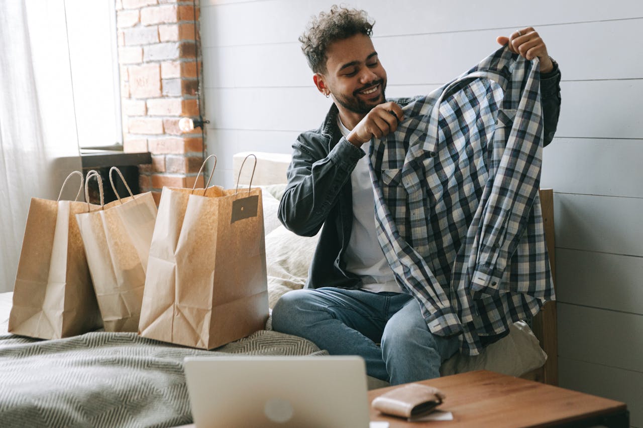 Smiling man examines checkered shirt indoors with shopping bags and laptop.